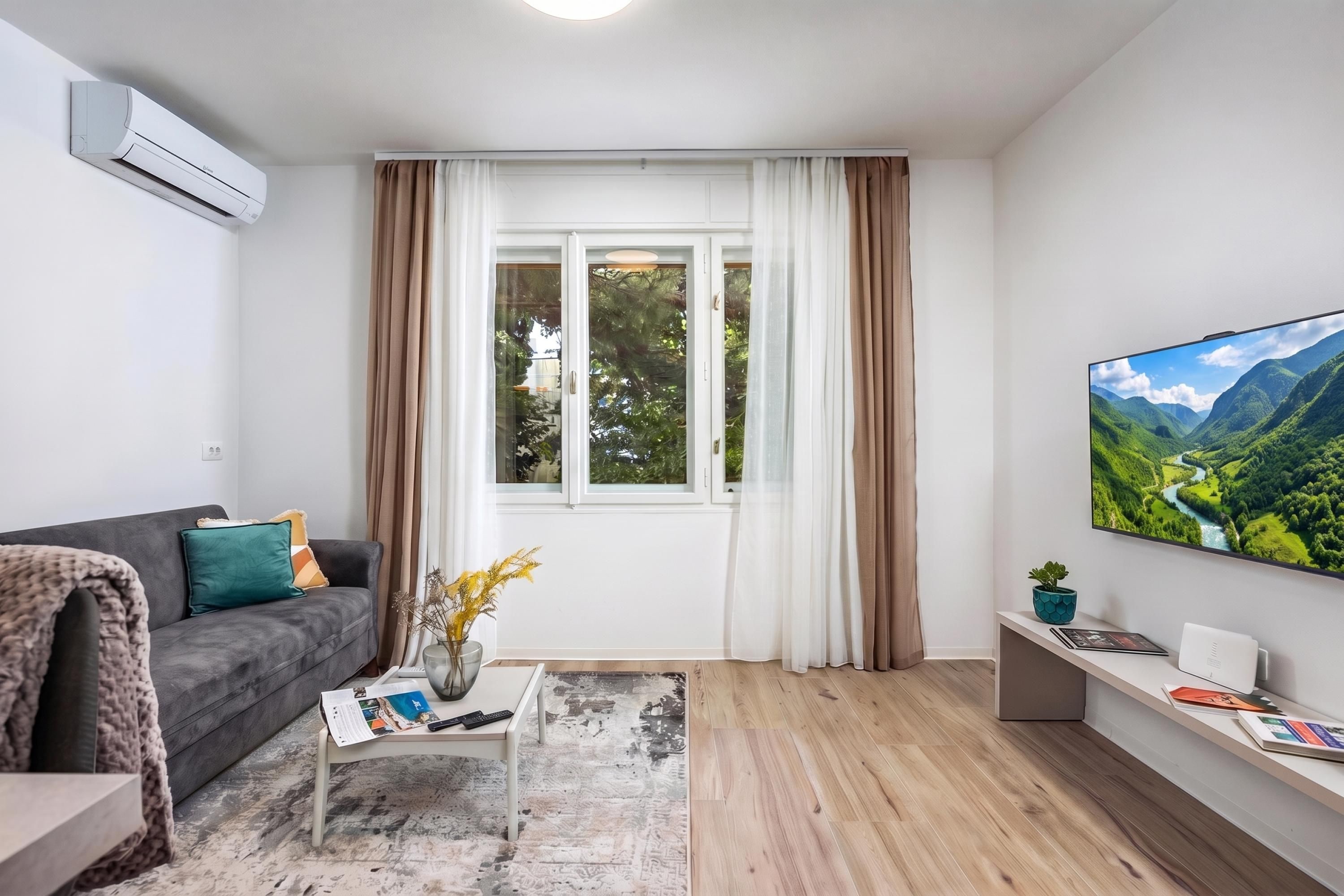 A modern living room with a grey sofa, a coffee table with a magazine, a TV on the wall, and a window with curtains.
