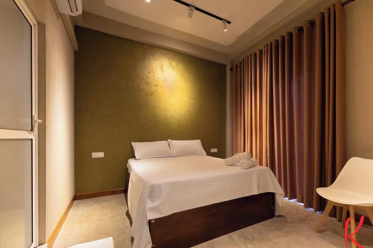 A bedroom with a bed, a chair, a curtain, and a wall with a light fixture.