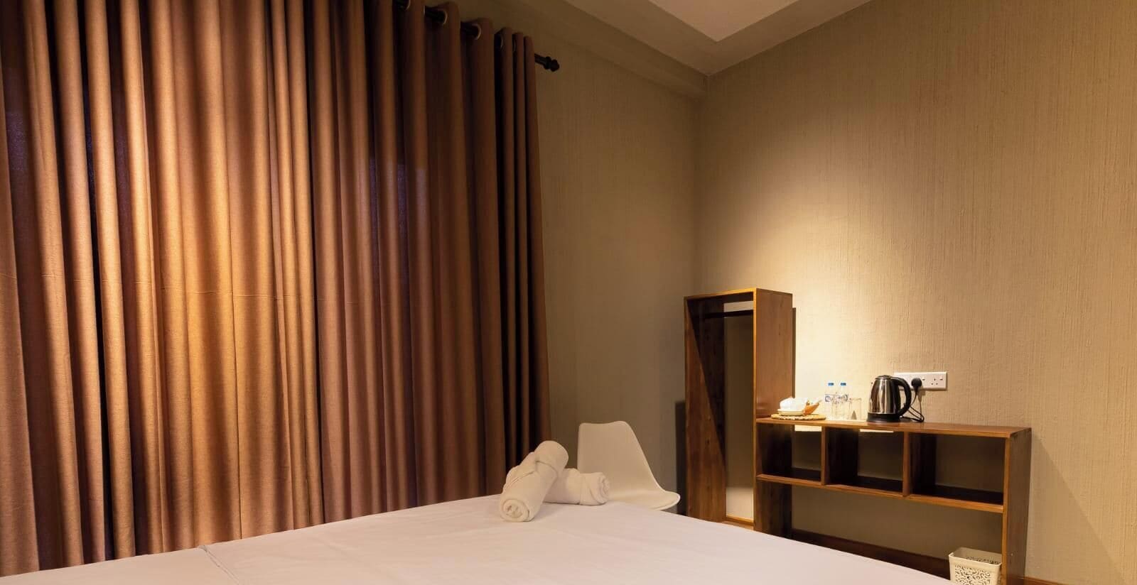 Habitación de hotel con cama, cortinas, una estantería de madera con una tetera y una mesita con una botella y un vaso.