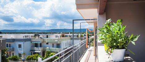 A balcony with a potted plant, a railing, and a view of buildings and mountains in the distance.