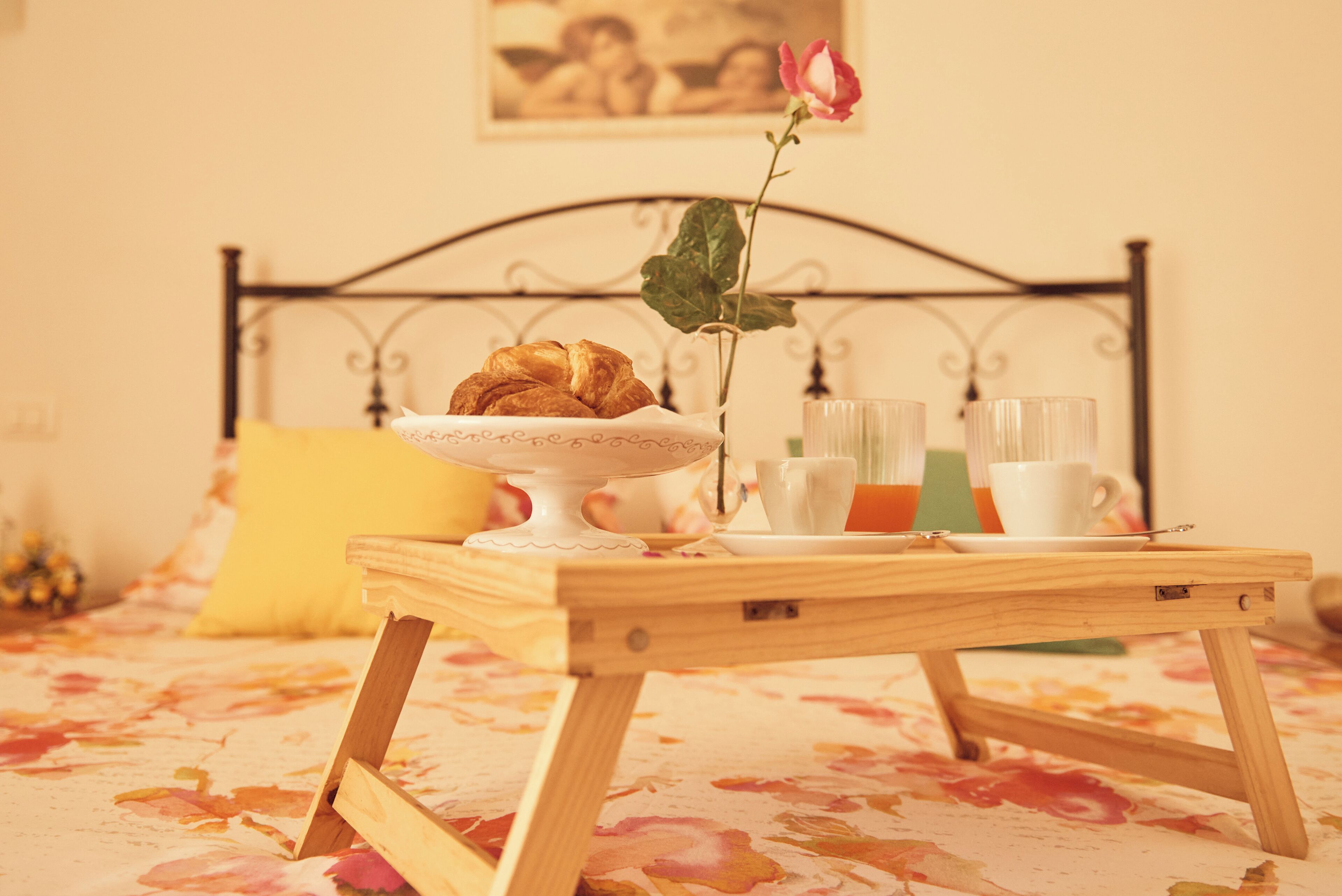 Una bandeja de madera con croissants, un florero con una rosa y una taza de café sobre una cama con colcha estampada.
