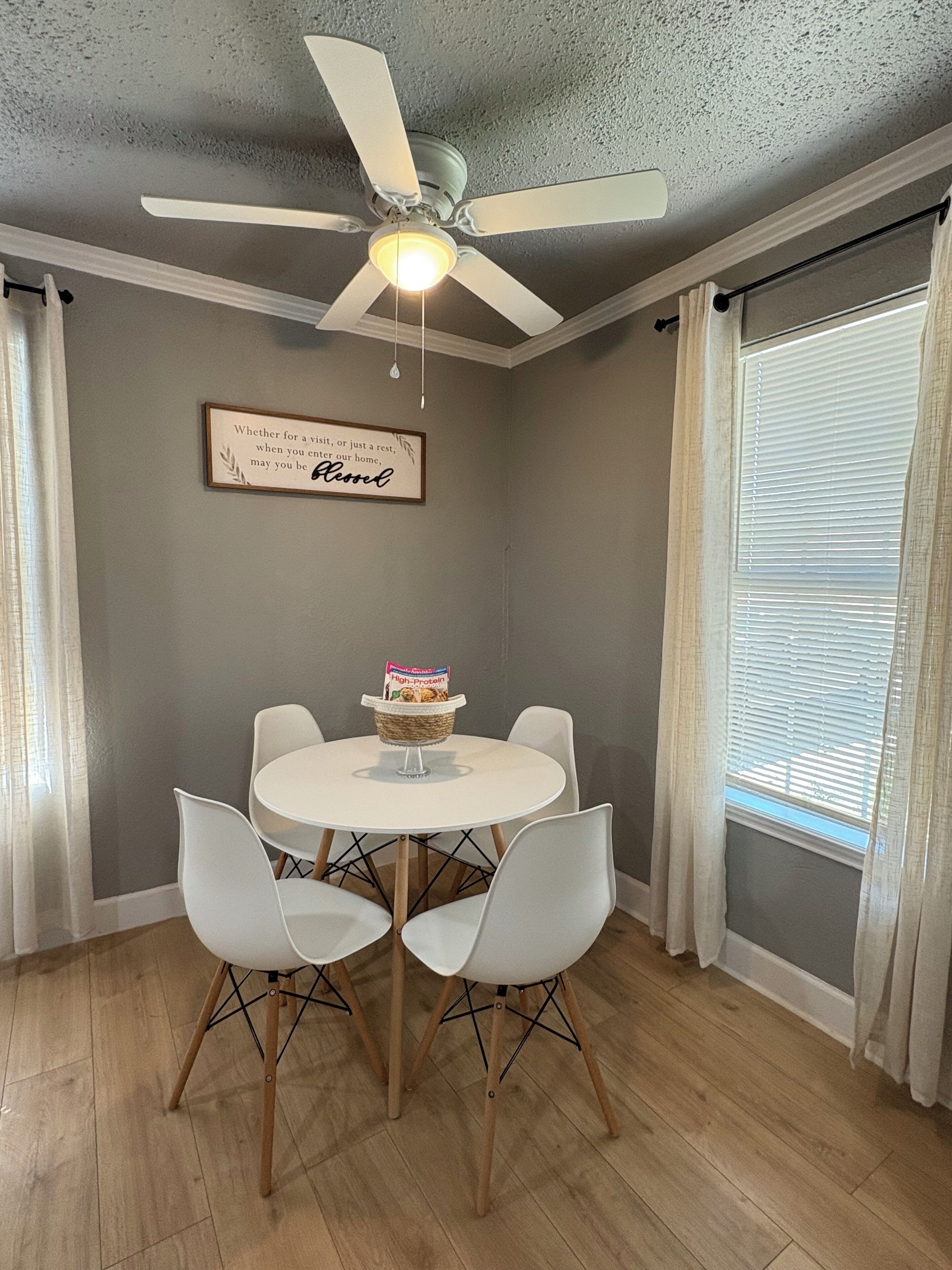 Un coin repas avec une table et des chaises blanches, un ventilateur de plafond, une citation encadrée accrochée au mur et une fenêtre avec des stores.