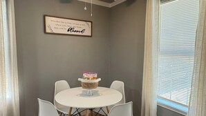 Un comedor con una mesa y sillas blancas, un ventilador de techo, una cita enmarcada en la pared y una ventana con persianas.