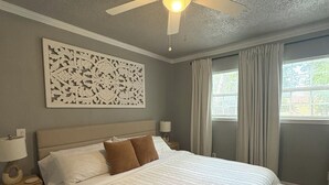 Un dormitorio ordenado con una cama grande, un ventilador de techo, un adorno en la pared y una ventana con cortinas.