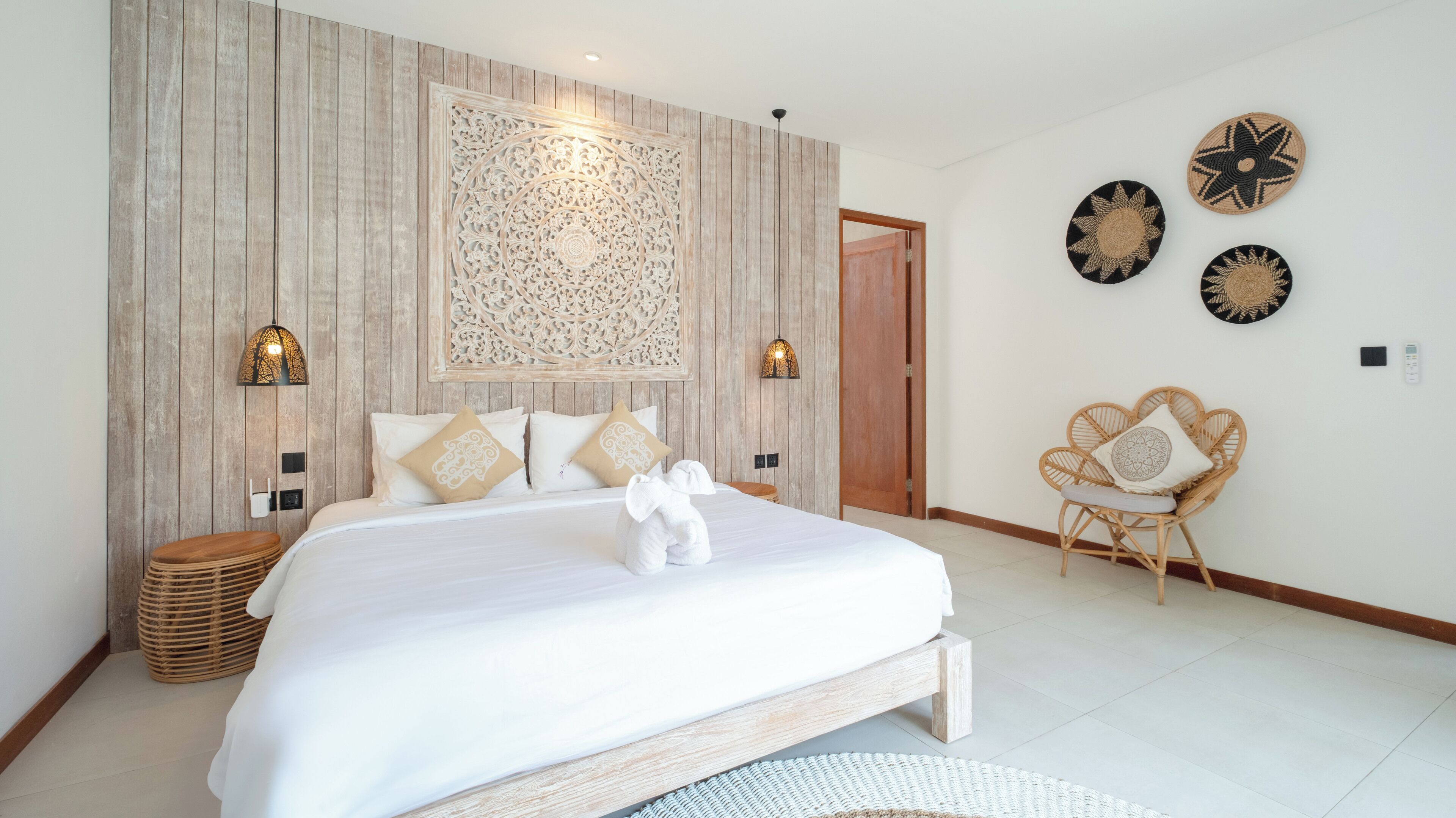 A bedroom with a large bed, two wall-mounted lamps, a decorative wall panel, a wicker chair, and a small table.