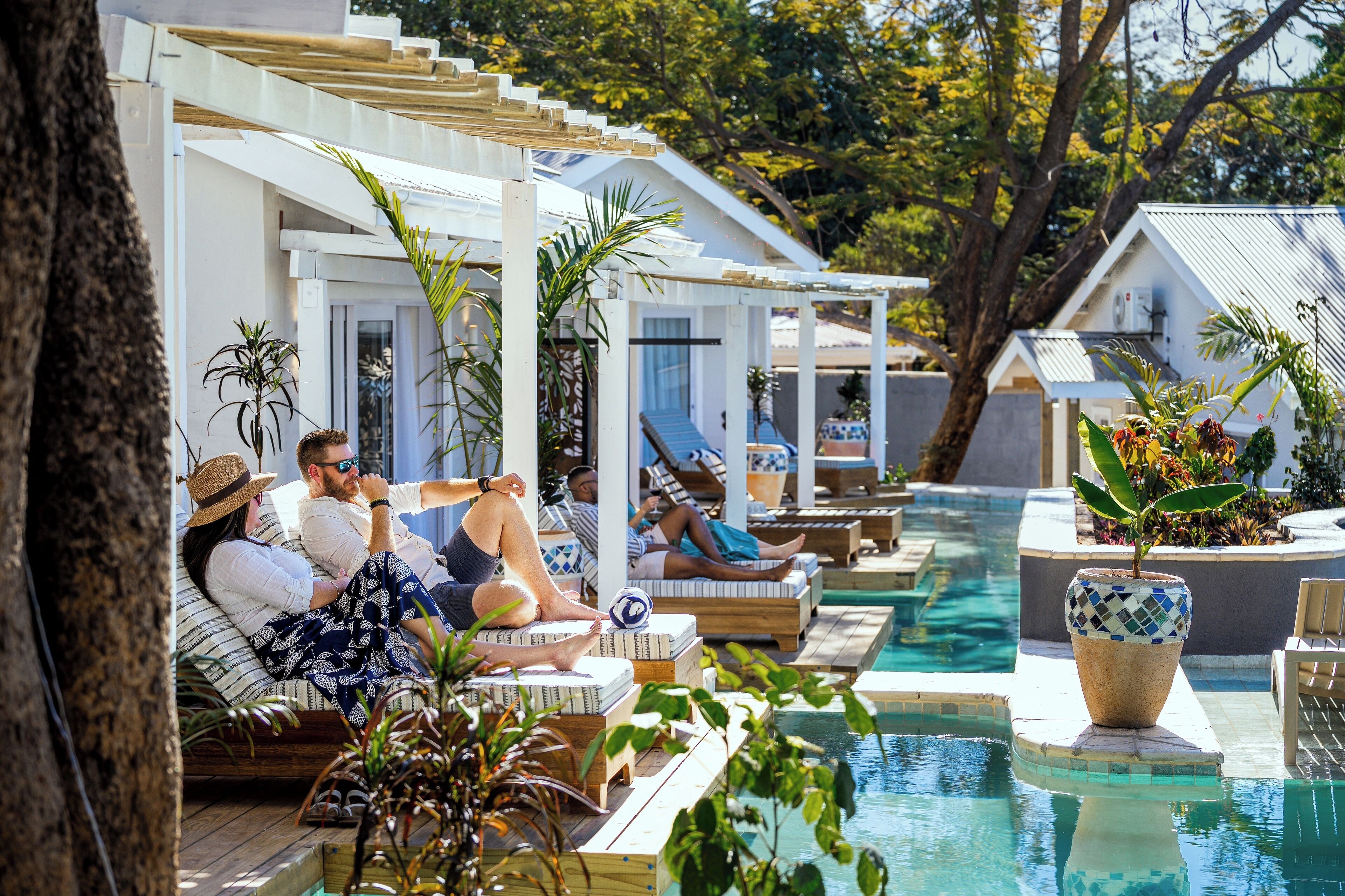 A poolside area with lounge chairs and a swimming pool, surrounded by greenery and white structures.