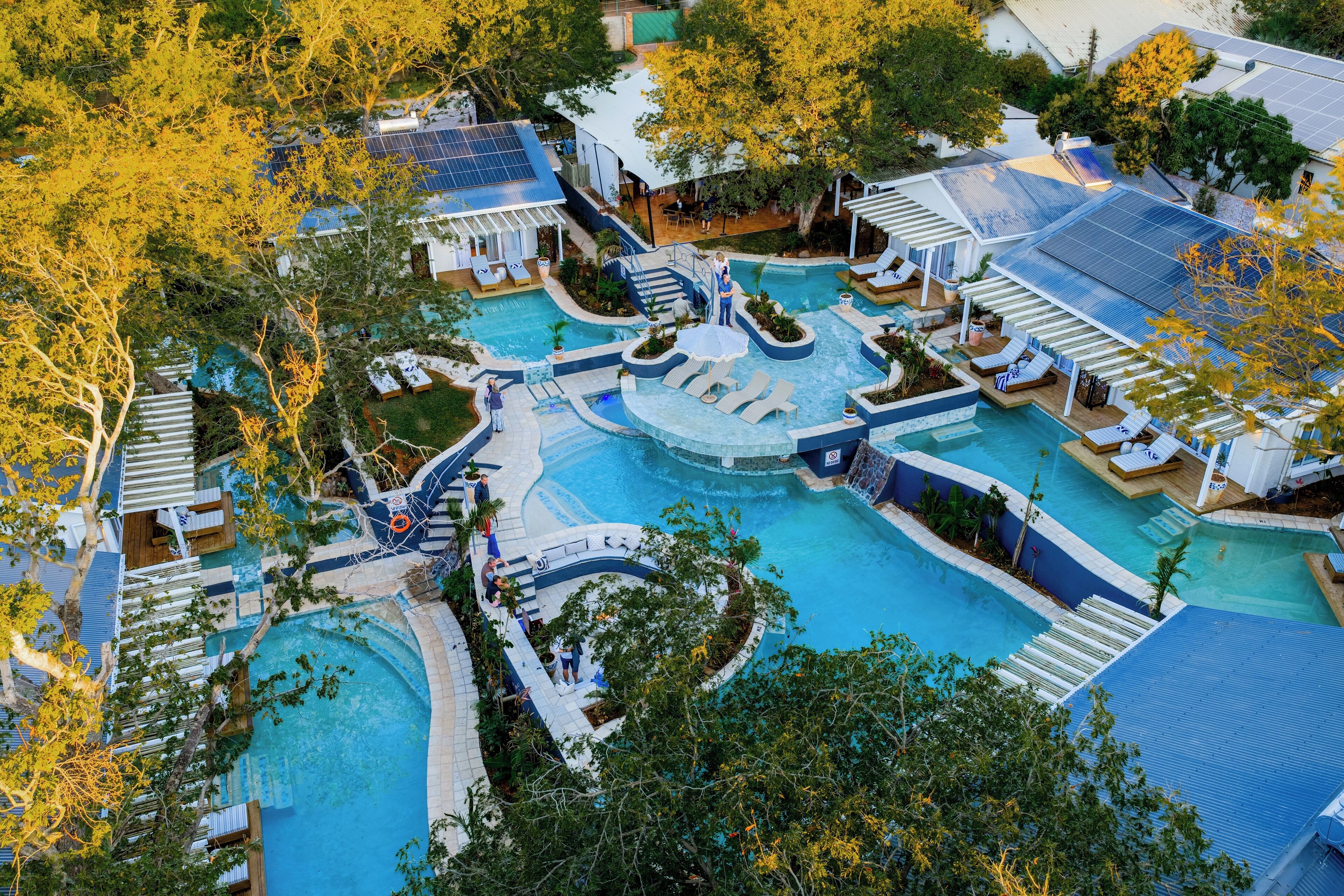 A large outdoor swimming pool with multiple sections and a central water feature, surrounded by lounge areas and covered seating.