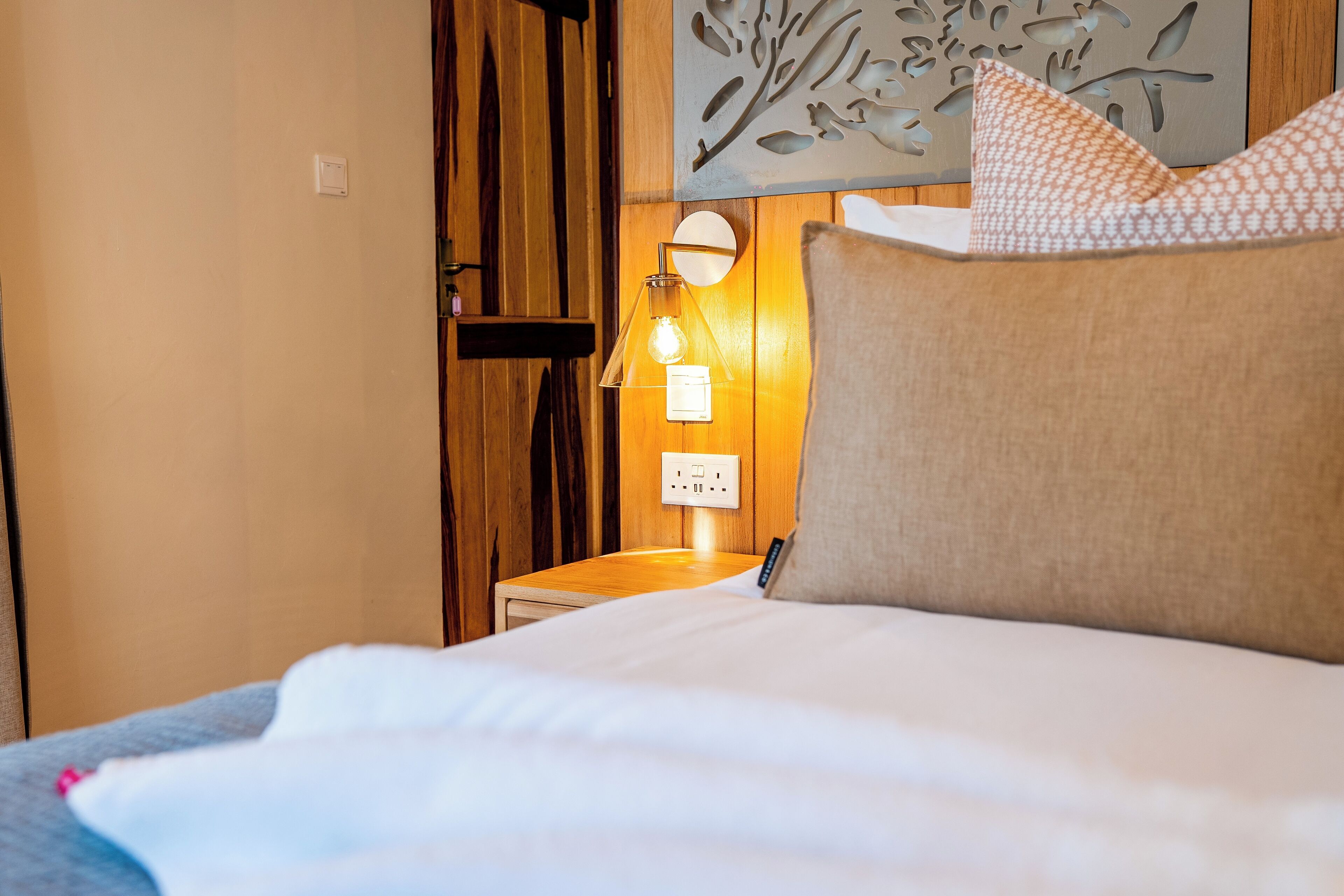 A neatly made bed with a white comforter and a beige pillow, a bedside lamp, and a wall-mounted power outlet.