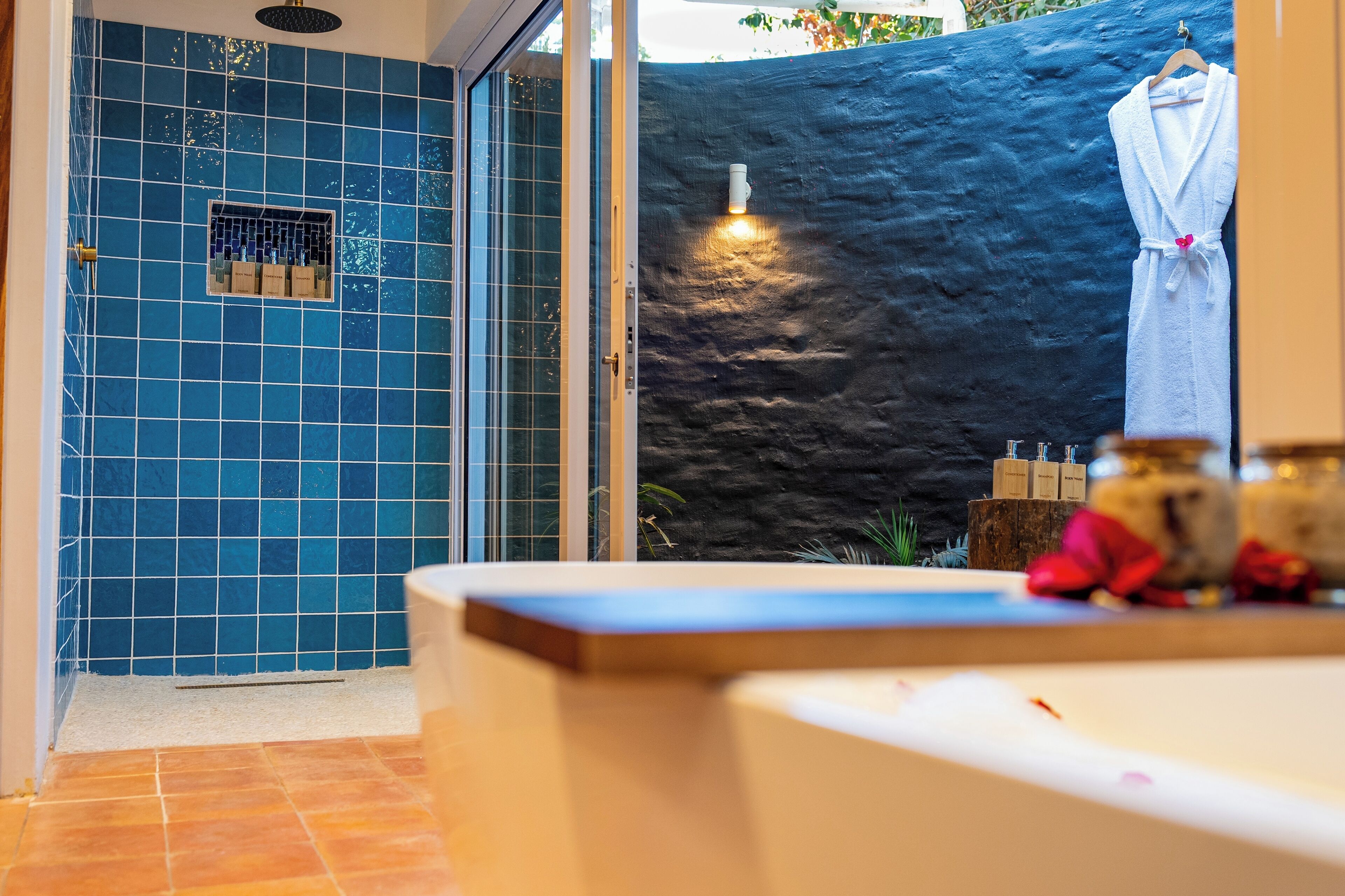 A bathroom with a blue tiled wall, a shower area, a towel rack, and a sink with toiletries.