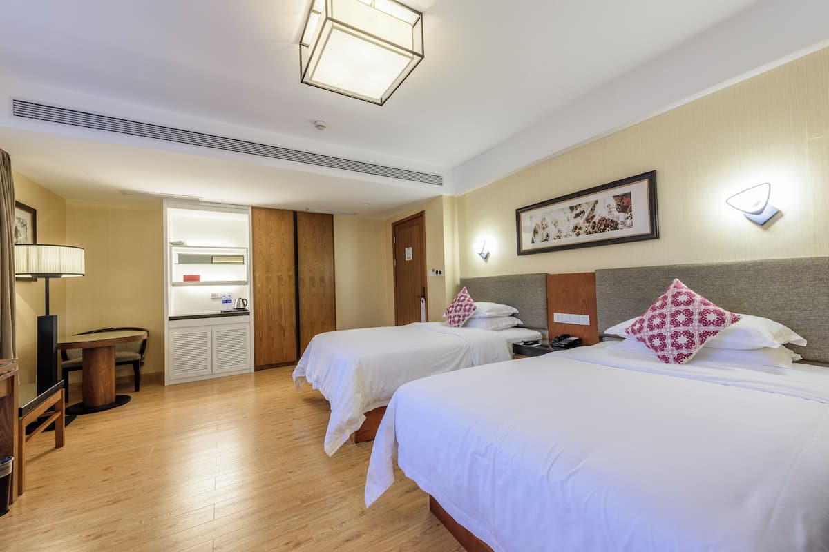 A hotel room with two beds, a wooden floor, a desk with a chair, a small kitchenette, and a framed picture on the wall.
