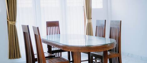 Une table ronde en verre, entourée de chaises en bois, se trouve dans une pièce lumineuse aux murs blancs et dotée d’une grande fenêtre.