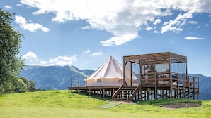 Una cabaña de madera con una estructura tipo tienda de campaña en una colina cubierta de hierba, con vistas a un paisaje montañoso.