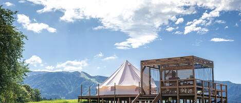 A wooden cabin with a tent-like structure on a grassy hill, overlooking a mountainous landscape.