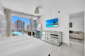 Habitación de hotel con una cama grande, un televisor, un aparador de vidrio y vistas a una piscina y edificios.