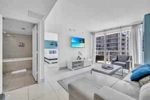 A modern living room with a sofa, a coffee table, and a TV. There is a balcony with a view of buildings.