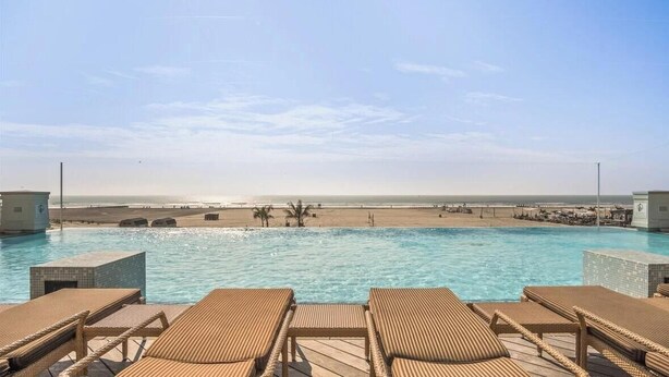 Ein Pool mit Liegestühlen, Blick auf den Strand und einem wolkenlosen Himmel.