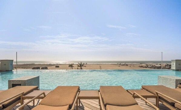A pool with lounge chairs, a beach view, and a clear sky.