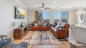 Ein modernes Wohnzimmer mit einer Couch, einem Couchtisch und einem Teppich. Große Fenster und ein Deckenventilator sind vorhanden.