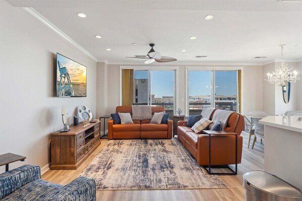 A modern living room with a sofa, a coffee table, and a rug. There are large windows and a ceiling fan.