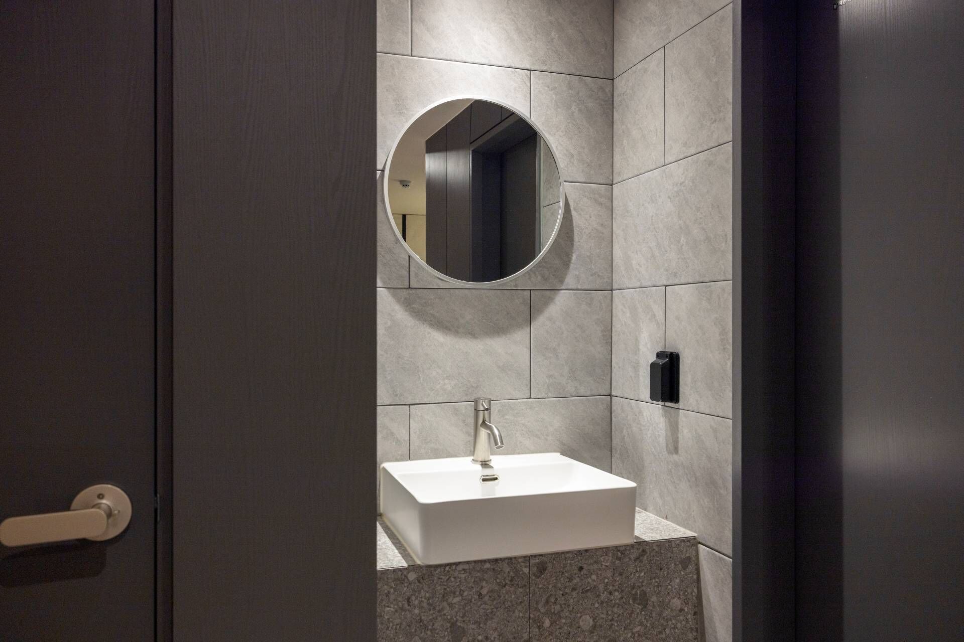 Un baño moderno con un lavamanos blanco, un espejo redondo y una puerta oscura.