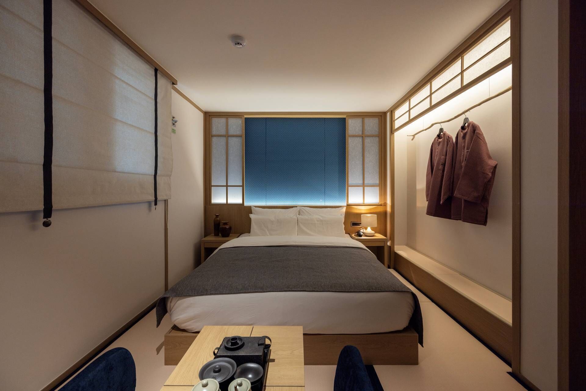 A compact hotel room with a bed, bedside table, lamp, and a small table with speakers.
