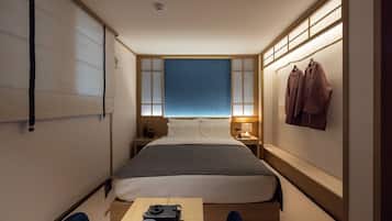 A compact hotel room with a bed, bedside table, lamp, and a small table with speakers.