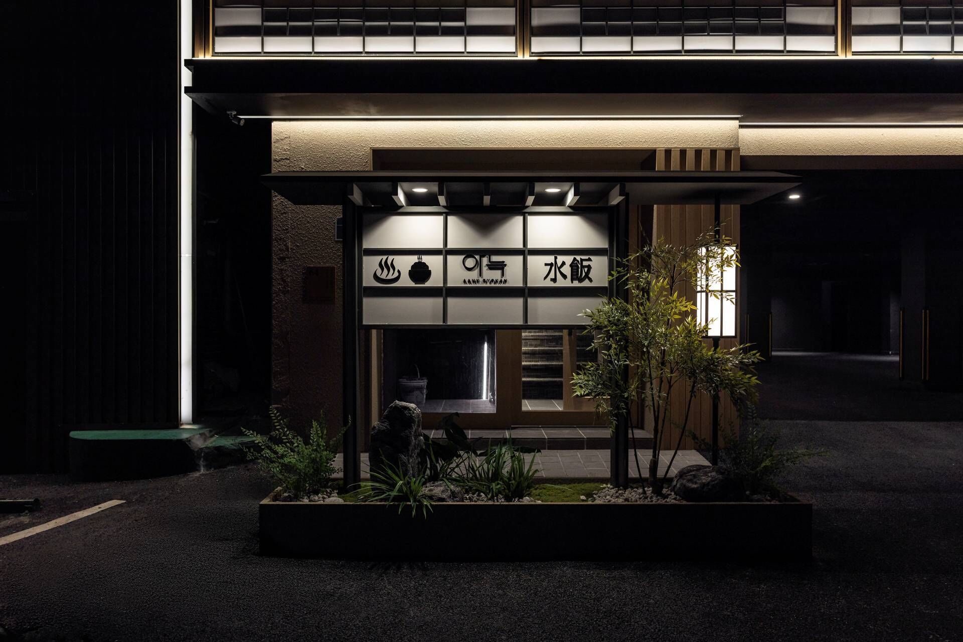 A building with a signboard displaying Japanese characters, a small garden with plants, and a well-lit entrance.