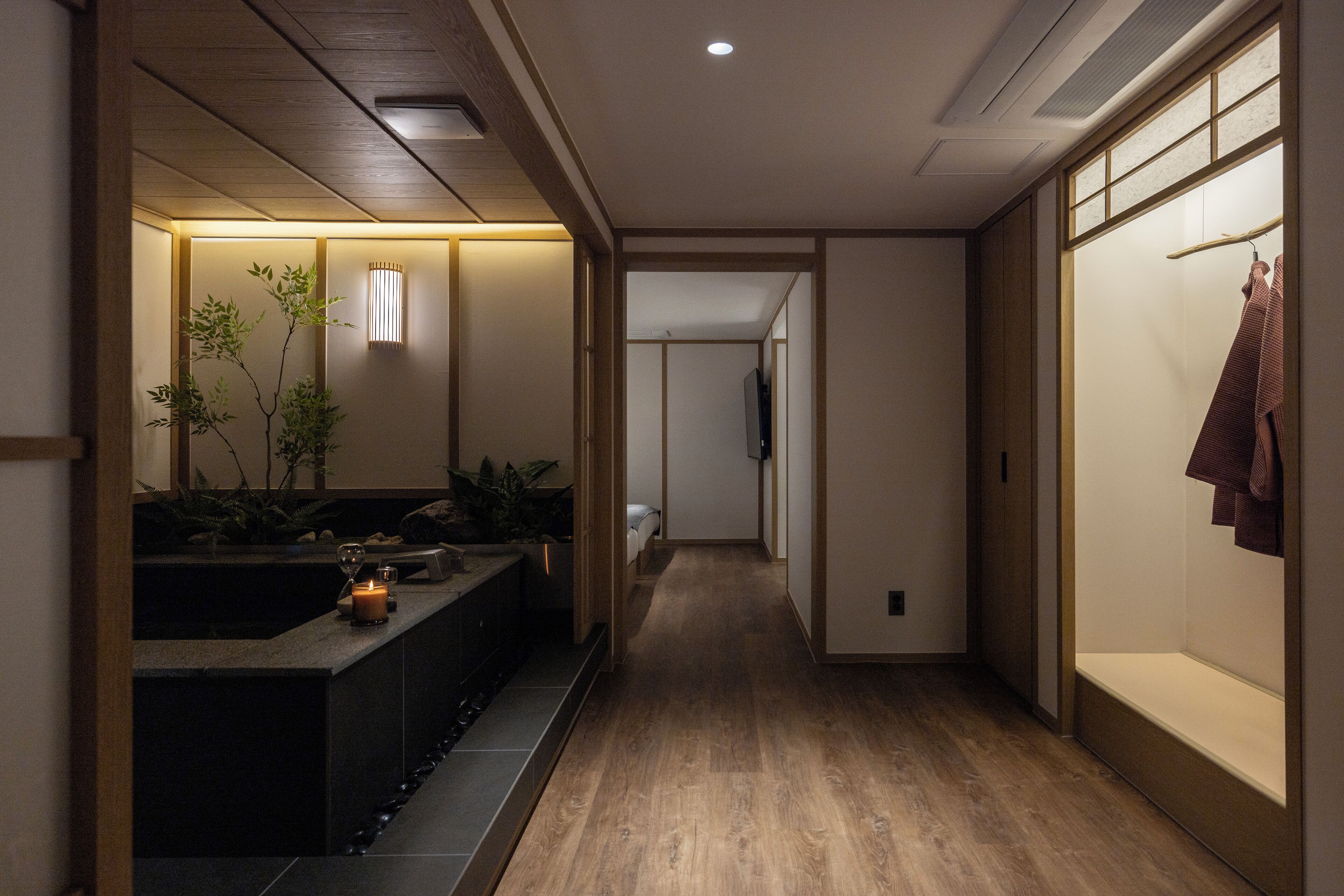 A hallway with a wooden floor, a reception area with a counter and plants, and a TV mounted on the wall.