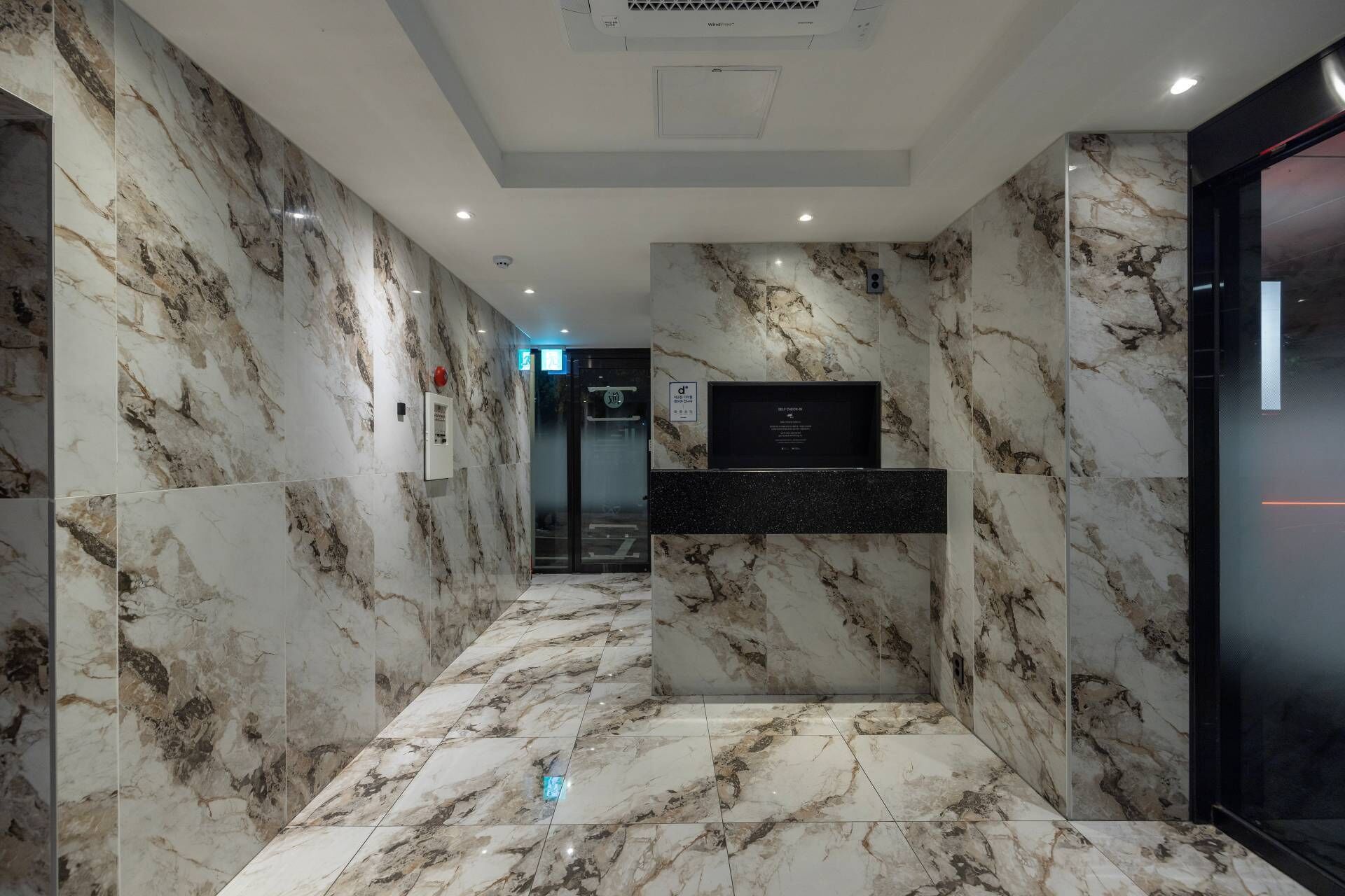 A modern hallway with marble walls and floors, a glass elevator, and a reception desk.
