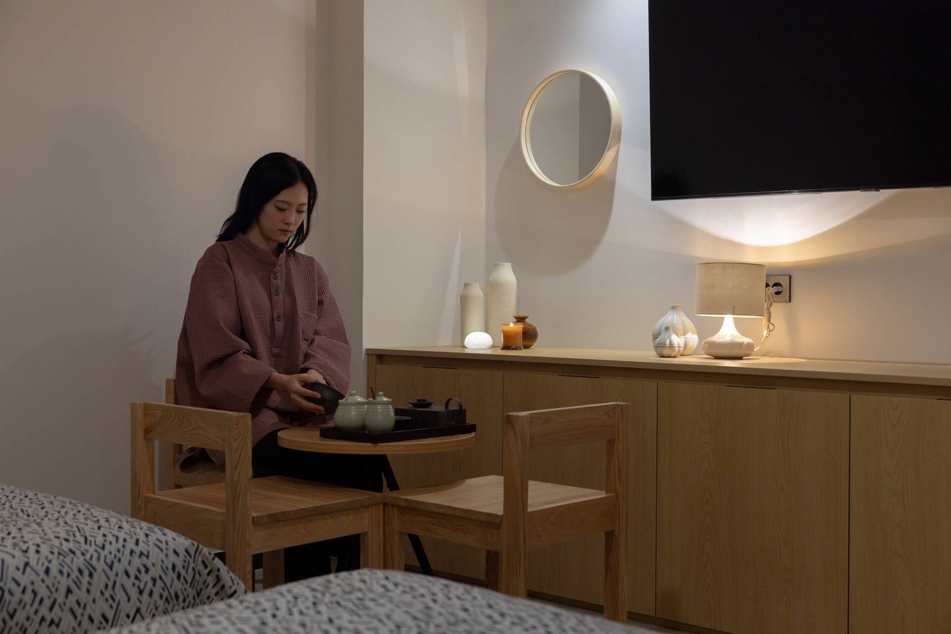 A person sitting at a wooden table with a laptop, a candle, and a lamp on a bedside table.