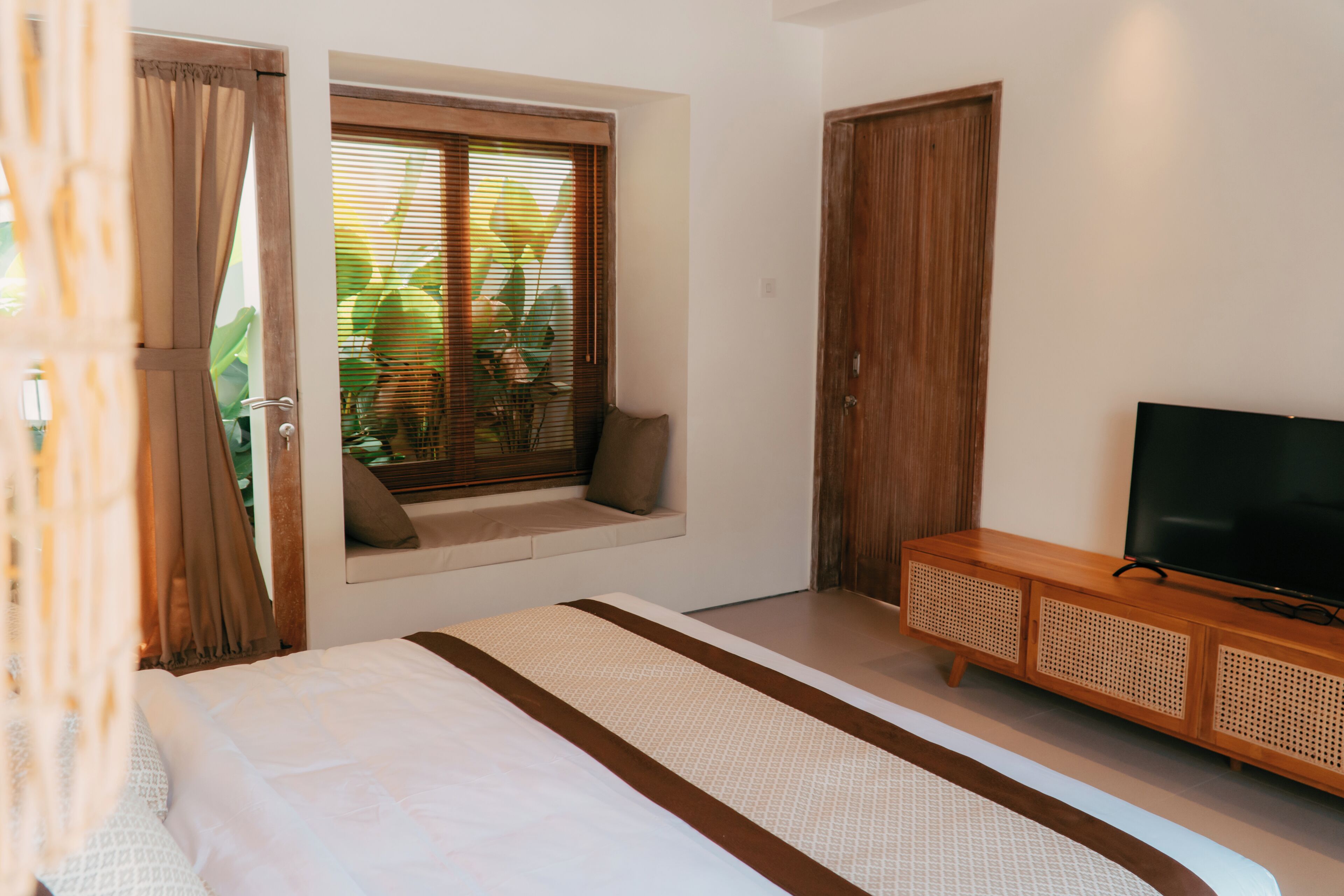 A bedroom with a bed, a television on a wooden cabinet, a window with blinds, and a door.