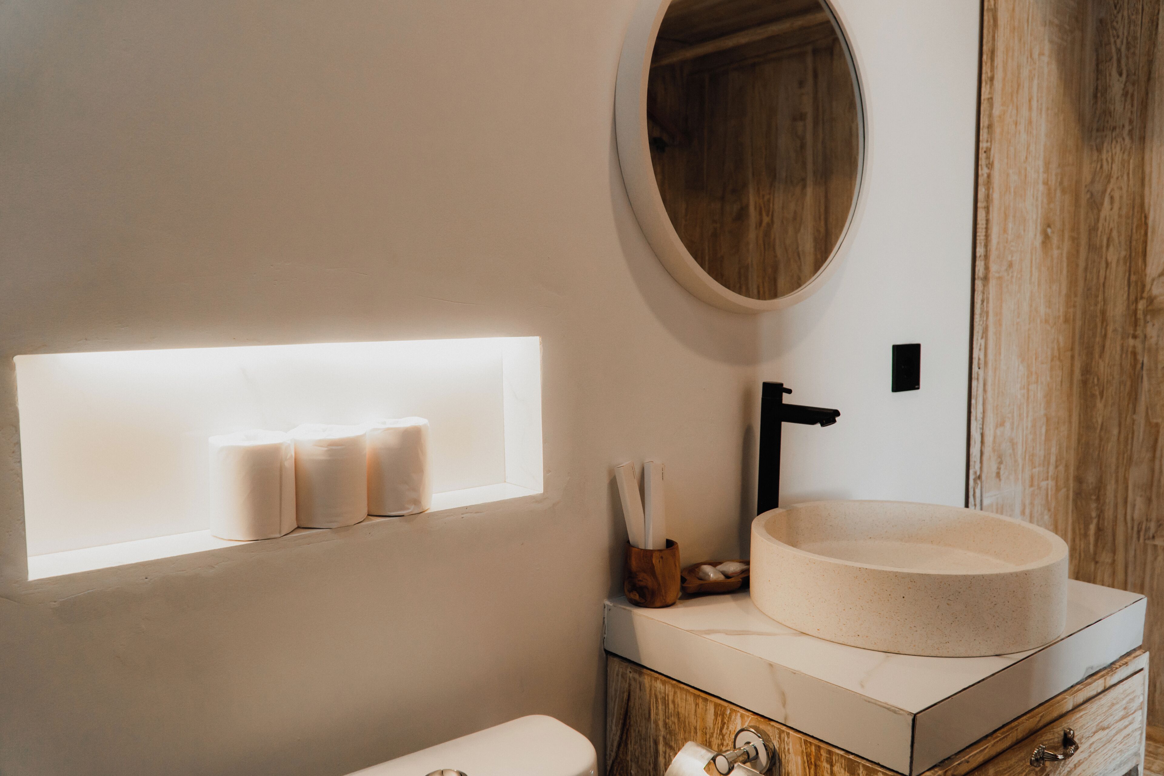 A modern bathroom with a round sink, wooden vanity, and a built-in shelf with toilet paper.
