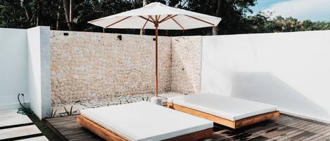 Two beds with wooden frames and white mattresses on a wooden deck, an umbrella, and a stone wall.