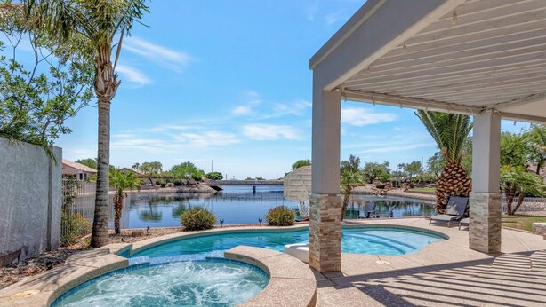 A swimming pool area with a hot tub, a covered patio, and a view of a lake.
