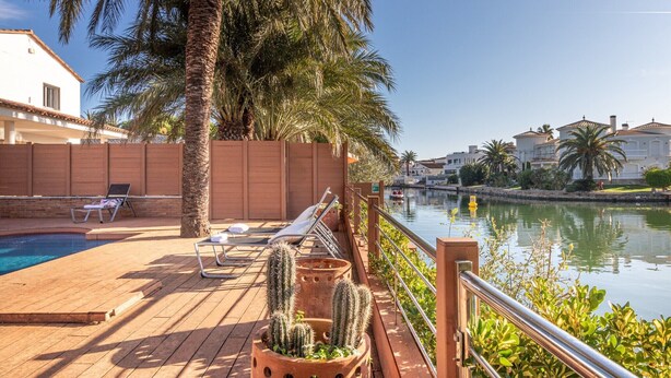 A poolside area with lounge chairs, a palm tree, and a view of a calm body of water. - Villa in Empuriabrava with Private Pool & Mooring (Empuriabrava)