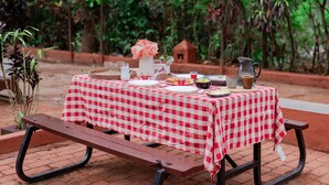 Une table de pique-nique installée à l’extérieur, recouverte d’une nappe à carreaux, et entourée d’une végétation luxuriante et d’arbres.