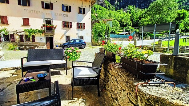 Un área de estar al aire libre con sillas y mesas de mimbre, un muro de piedra con macetas, y un edificio con un letrero que dice "TRATTORIA IN LLOCCIO".