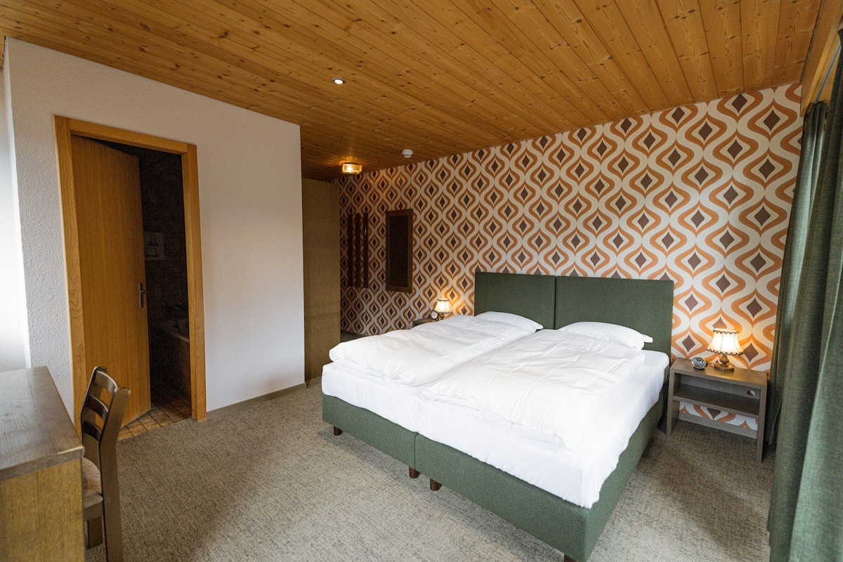 Ein Schlafzimmer mit Holzboden, einer Wand mit Muster, einem Bett mit weißer Bettwäsche, einem Nachttisch mit Lampe und einem Holzstuhl.