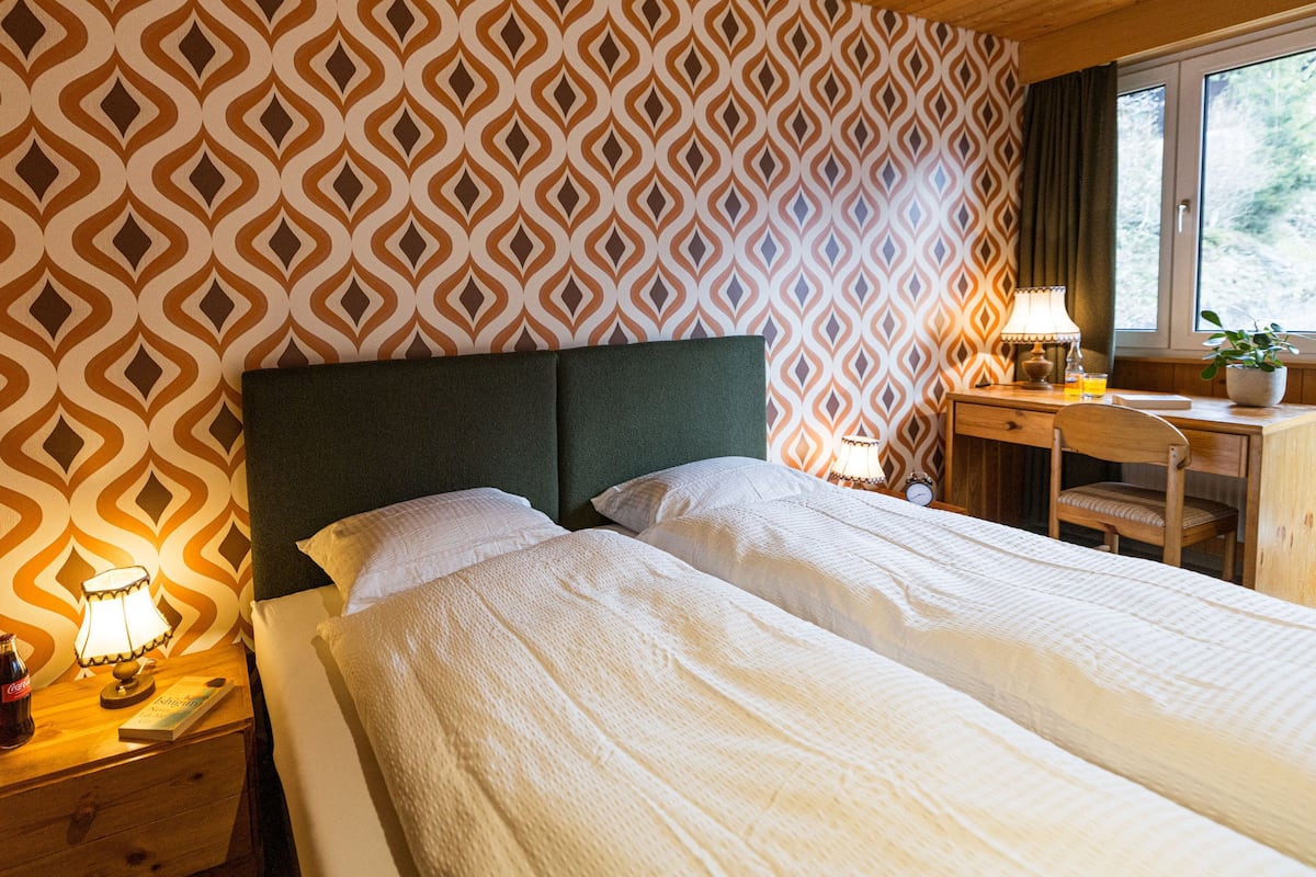 Ein Doppelbett mit grünem Kopfteil, ein Nachttisch mit Lampe, einer Uhr und einer Coca-Cola-Flasche.