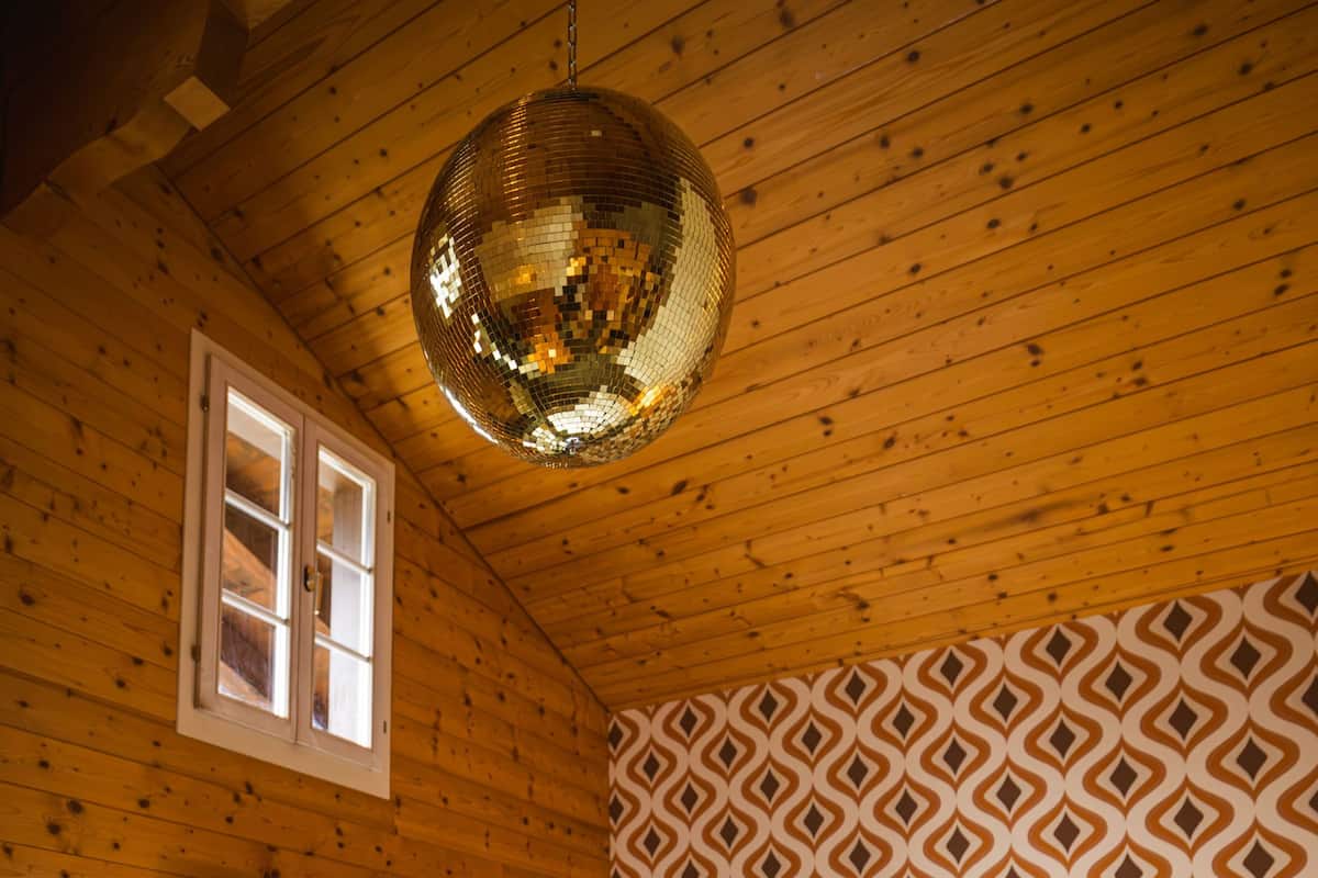 Ein Holzhüttenraum mit einem Bett, einem Fenster mit Vorhängen, einer Wand mit Muster und einer Discokugel.