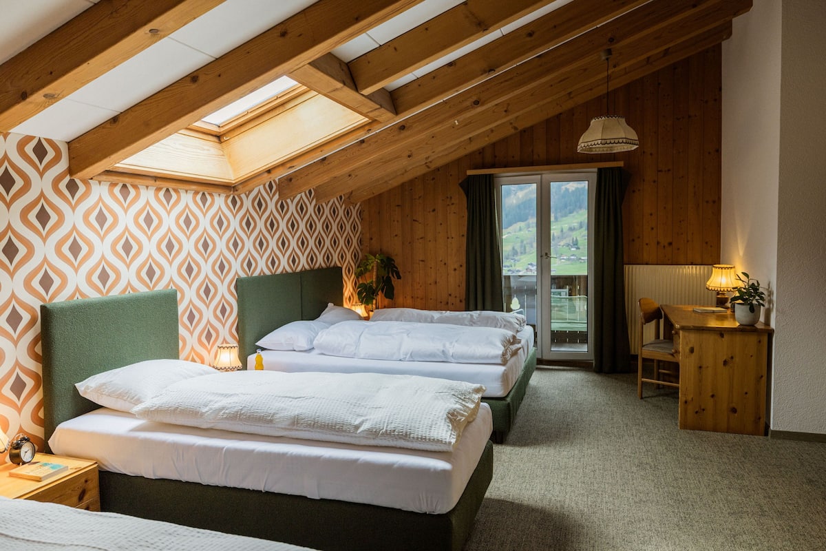 Ein Schlafzimmer mit zwei Betten, einem Oberlicht, Holzbalken, Blick ins Freie und einem kleinen Tisch mit Lampe.