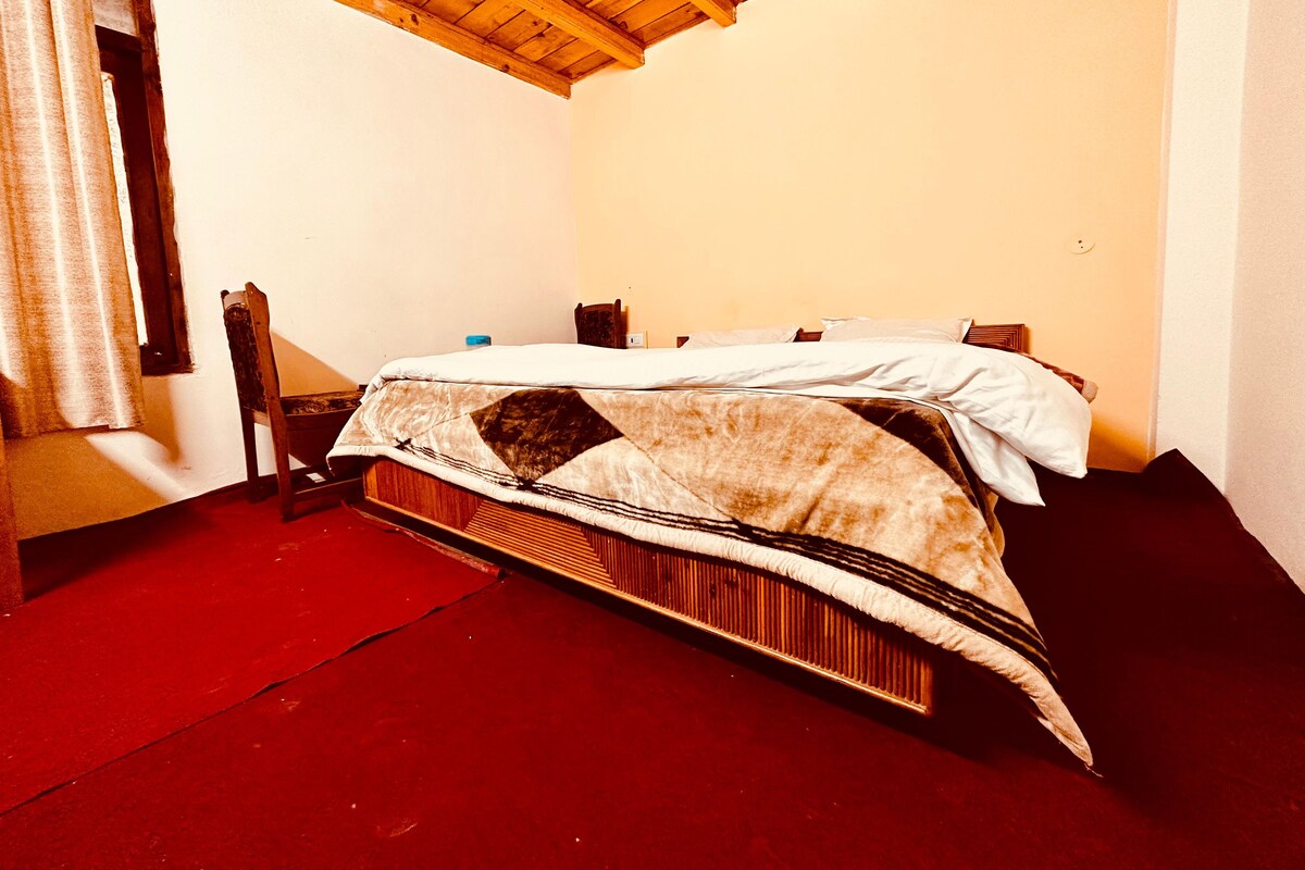 A single bed with a patterned bedspread, a wooden chair, and a red carpet in a room with white walls and a wooden ceiling.
