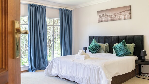 A bedroom with a large bed, blue curtains, a cityscape painting, and a nightstand with a lamp. - Grand Manor  (Muldersdrift)