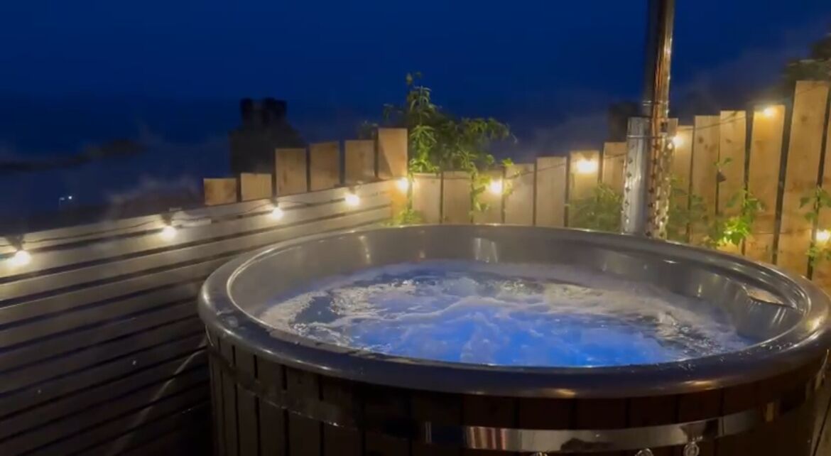 A hot tub on a wooden deck with a view of the city skyline at night.