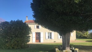 Una casa con techo de tejas, puerta de madera y contraventanas blancas. Hay un árbol en primer plano y un jardín césped al fondo.