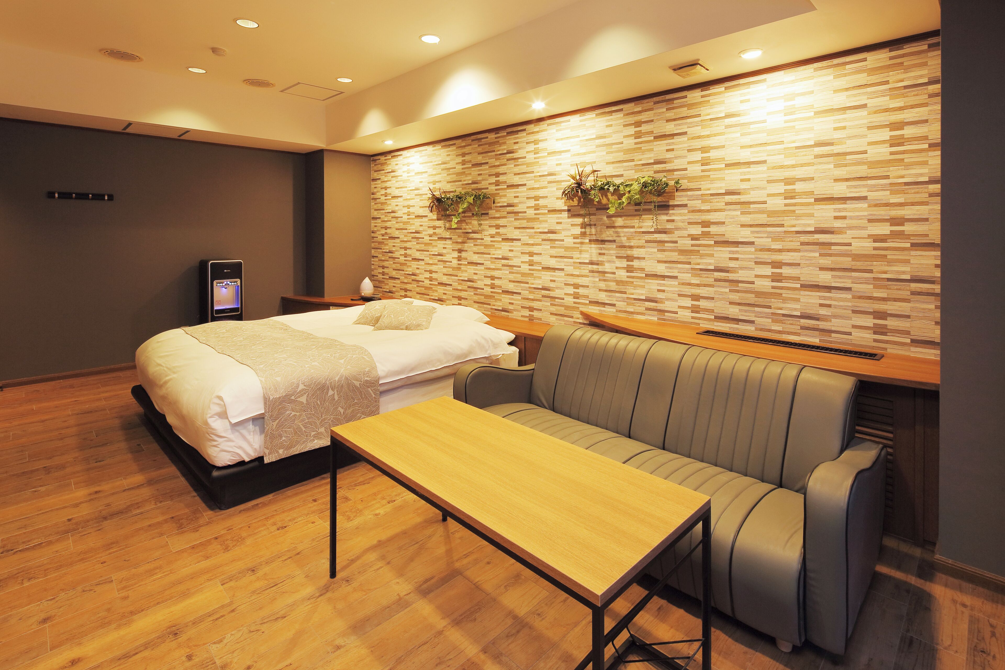 A modern hotel room with a bed, a sofa, a wooden table, and a brick-patterned wall with plants.