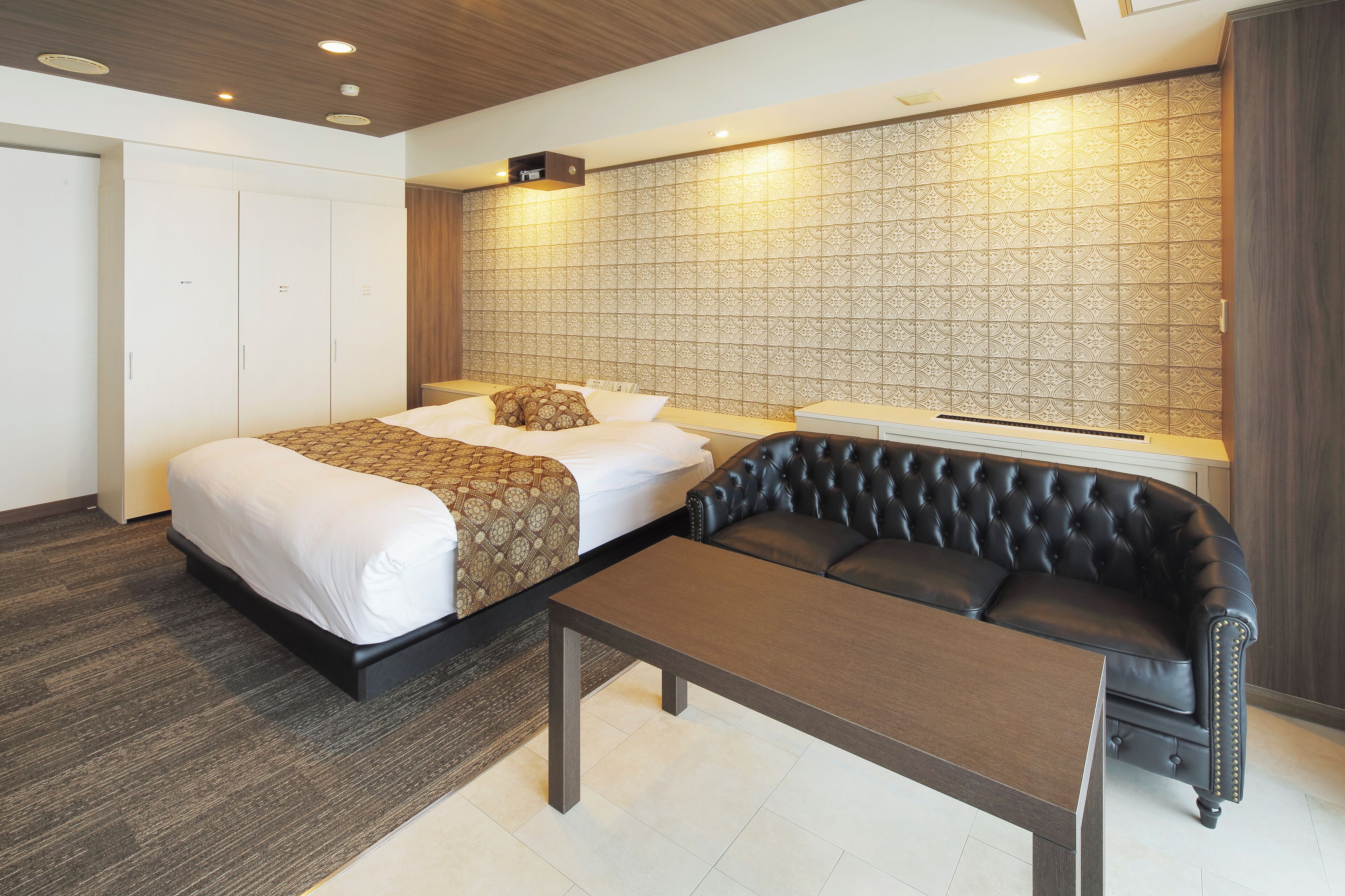 A hotel room with a bed, a black leather sofa, a wooden table, and a patterned wall.