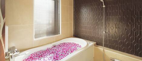 Un baño con una bañera llena de pétalos de rosa, ducha y ventana con persianas.