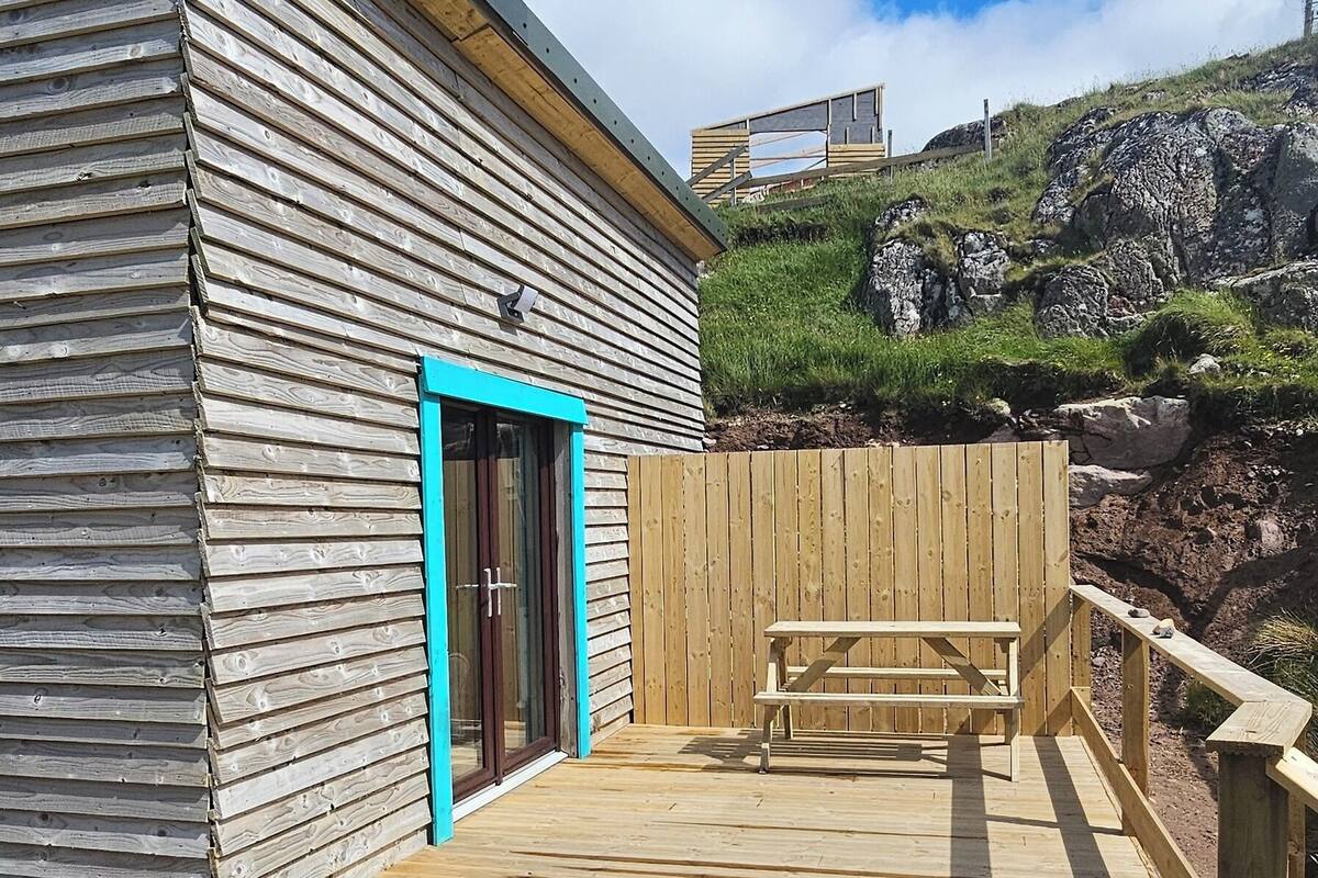 A wooden deck with a bench, a building with a blue door, and a rocky hillside in the background.
