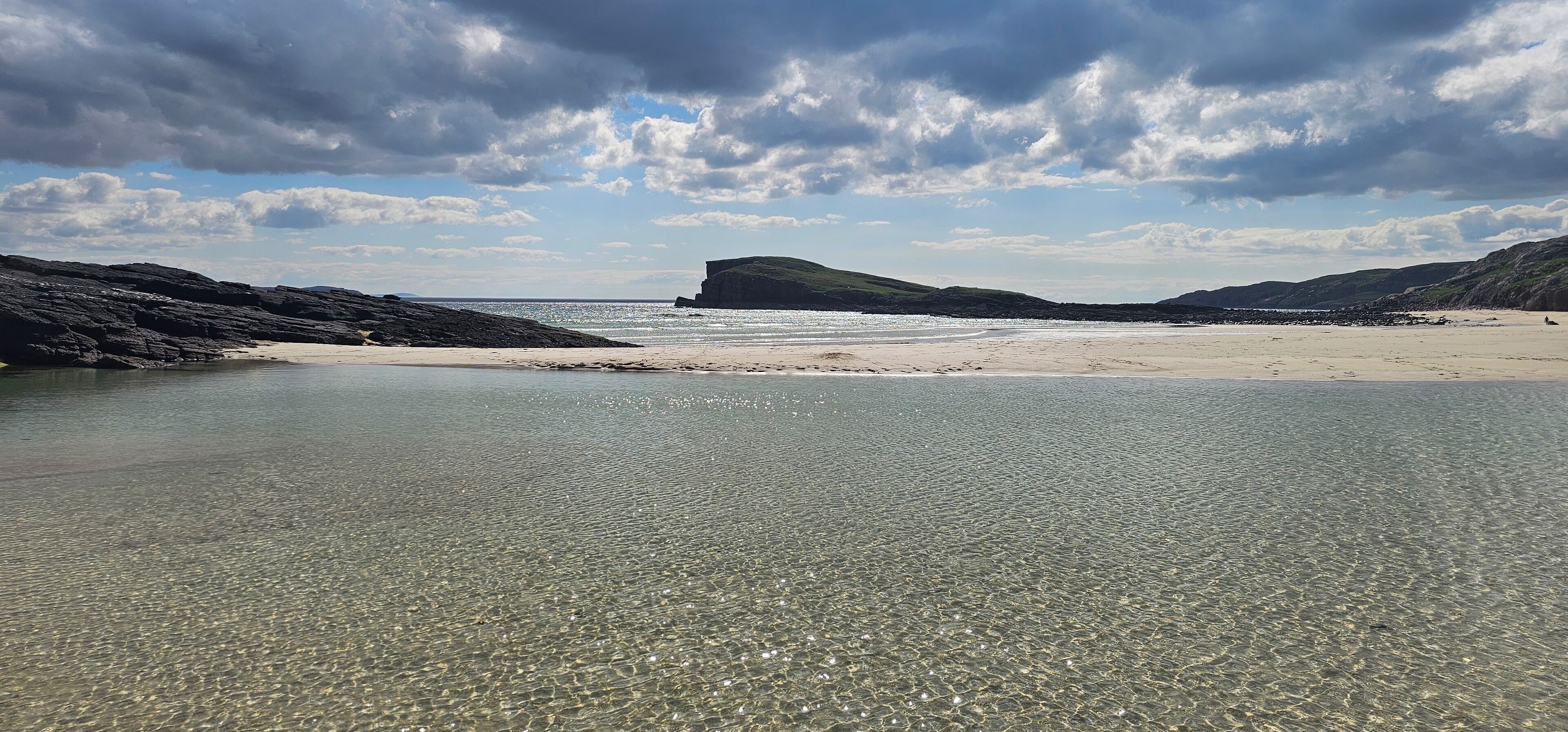 Una playa con aguas claras y poco profundas, orilla de arena y acantilados rocosos al fondo.